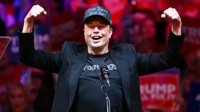 Elon Musk, vestido de negro, eleva sus brazos en señal de triunfo durante un acto político de Trump en octubre 