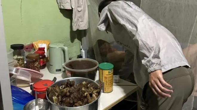 Pessoa se aproxima para falar com trabalhador deitado em uma cama coberta por um mosquiteiro e próxima a uma mesa cheia de comida, com uma panela com carne