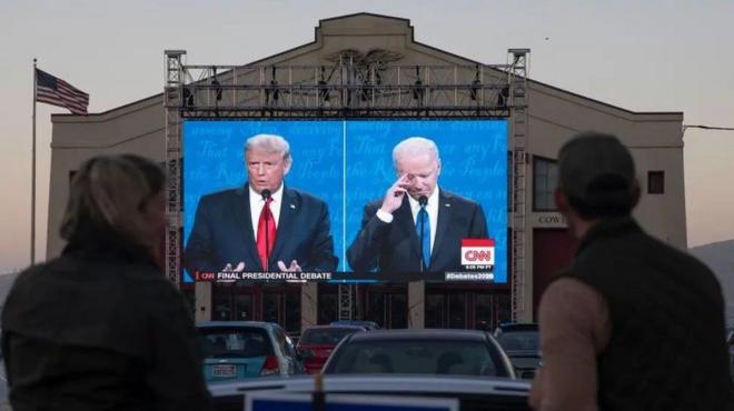 Espectadores nas ruas assistem a um debate presidencial de 2020 entre Joe Biden e Donald Trump, sendo transmitido em telão