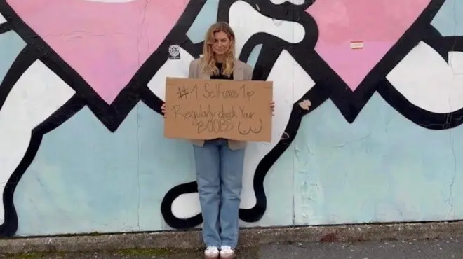 Barbora is standing in front of a wall mural on Tyneside and holding a sign that reads "selfcare tip: regularly check your boobs." She has long blonde hair and is wearing jeans and trainers. 