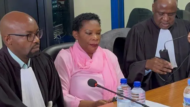 Béatrice Munyenyezi mu rukiko mu Rwanda muri uku kwezi ari hagati y'abunganizi be mu mategeko. Yambaye ikanzu y'iroza yo mu mwambaro uranga imfungwa, imbere hari ameza ariho indangururamajwi n'amacupa y'amazi. 