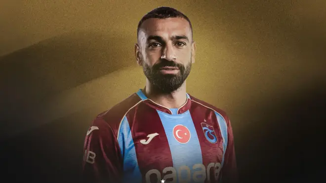 Siyah saçlı sakallı futbolcu bordo mavili bir formalı ile poz veriyor. Arka fon tamamen altın rengi