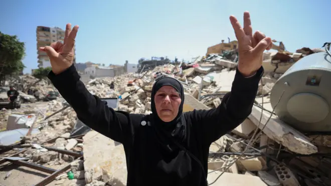 Una mujer libanesa de mediana edad, con vestidura islámica negra, hace la señal de la paz con dos manos en alto, frente a los escombros causados por los ataques israelíes en la ciudad de Tiro