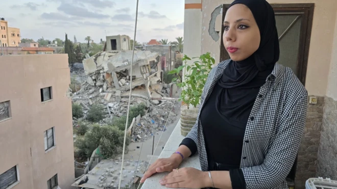 زنی محجبه در بالکنی خانه‌ای ایستاده به دور دست نگاه می‌کند، آنسوتر ویرانه‌های ساختمان‌ها دیده می‌شود