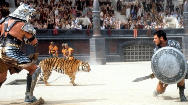 A scene from the movie Gladiator showing two men dressed in armour and leg and arm guards confronting a tiger in an arena with crowds of cheering people watching on. One of the men is wearing an ornate helmet while the other is holding a shield and a sword.