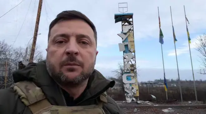 Скріншот з відео Зеленського: він знімає себе на камеру, стоячи у бронежилеті на тлі понівеченої обстрілом стели з написом "Купʼянськ". Поруч зі стелою – кілька потріпаних українських прапорів на високих флагштоках.