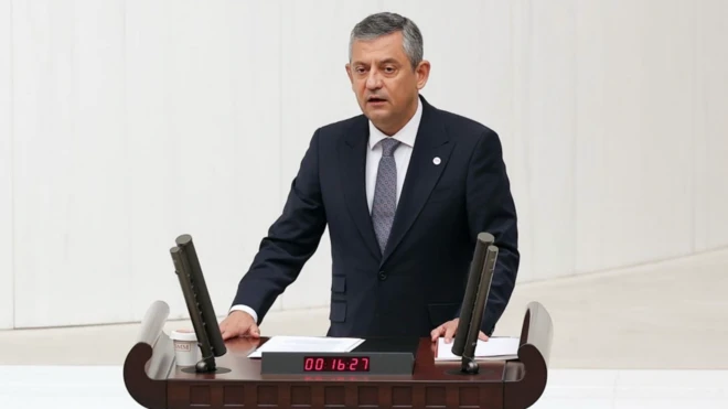 Özgür Özel elini kürsüye koymuş, mecliste konuşma yapıyor. Siyah bir takım giymiş ve karede bir tek o var.