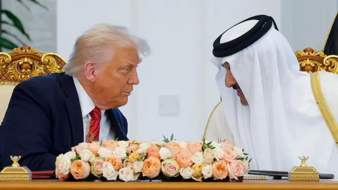 Trump y Al Thani se inclinan uno hacia el otro para conversar detrás de un escritorio dorado adornado con un ramo de flores blancas, naranjas y rosas.