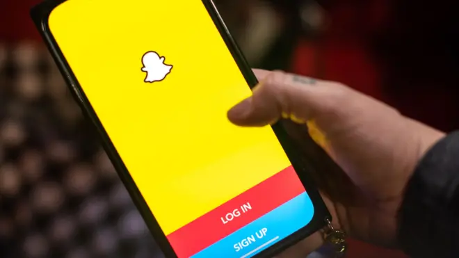 The snapchat logo is seen on a mobile phone icon with a thumb over the top of it about to tap the app icon.