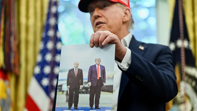 Трамп у Білому домі демонструє свою фотографію з Путіним, зроблену на Алясці. На тлі – американський прапор, а сам Трамп має на собі червону кепку