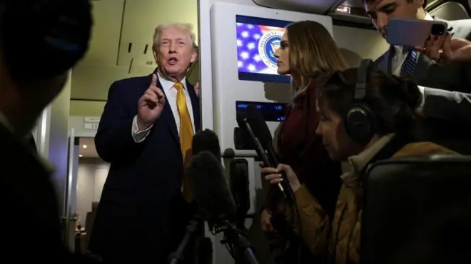 ダークスーツを着て金色のネクタイをしたトランプ氏が、右手を前に出し、人差し指を立てて口を開いている。その前には記者たちがいて、マイクを向けるなどしている