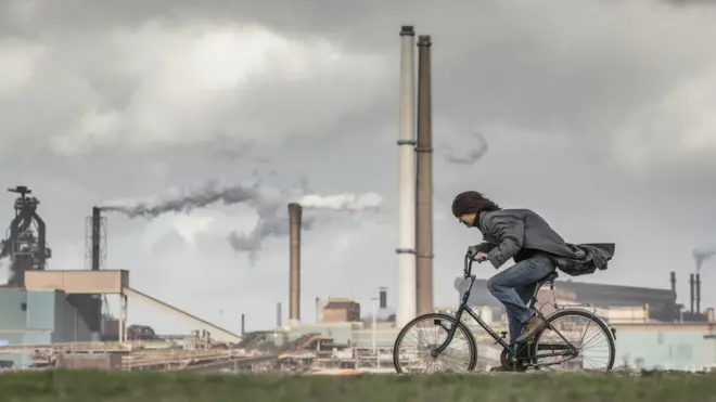 Tata Steel factory, blast furnaces. Cyclist, woman, tries to stay upright in storm