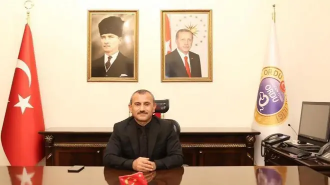 Siyah takım elbiseli bir adam makam odasında oturuyor ve yanında Türk bayrağı var.