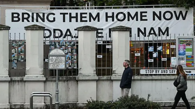 Dos personas caminan delante de un edificio en el que un gran cartel dice Bring Them Home ("tráiganlos a casa"), pidiendo el retorno de los rehenes que están en manos de Hamás