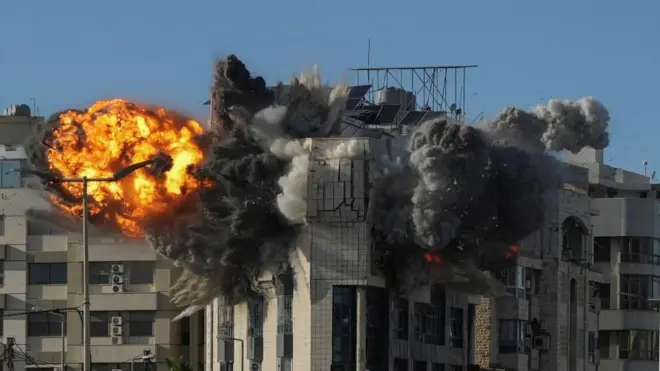 青空の下、クリーム色のタイル張りの建物が爆発し、オレンジ色の炎と黒煙が上がっている