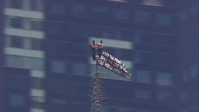 Two small figuress in black at the very top of the spire, with windows a building behind them, a black flag with white script waves to one side
