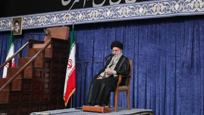 علی خامنه‌ای، روی صندلی و در کنار پرچم جمهوری اسلامی ایران و منبر