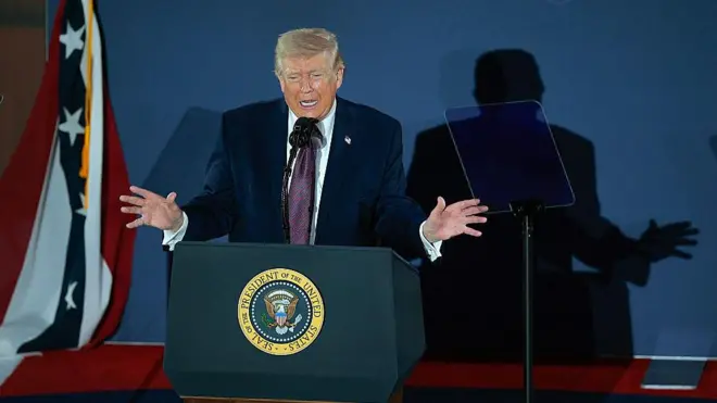 دونالد ترامپ پشت میکرفون در حال سخنرانی در حالی که دستانش را به دو طرف باز کرده، سایه او بر دیوار افتاده است