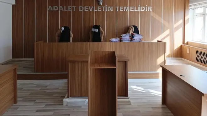 Boş bir mahkeme salonunda, mahkeme heyetinin yer aldığı kürsüde dosyaların yığıldığı görülüyor. 