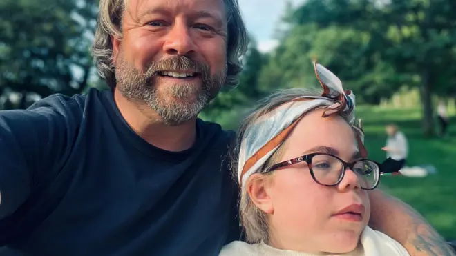 Darren and Sophia Scott. He has longish blond hair and a beard and is wearing a blue T‑shirt. His arm is around Sophia who is wearing glasses and a blue, brown and white cotton scarf tied around her head. He is smiling at the camera while she is looking off camera. They are in a park.