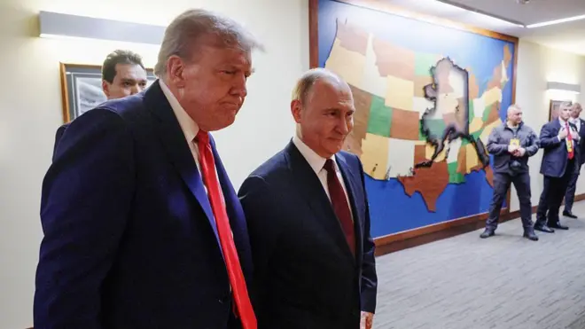 Трамп и Путин в коридоре на фоне карты штатов США, сзади видны люди с камерами.