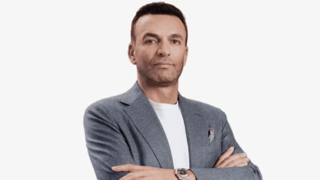 Ellerini önde kavuşturmuş bir adam poz veriyor, arka planda açık bir renk var