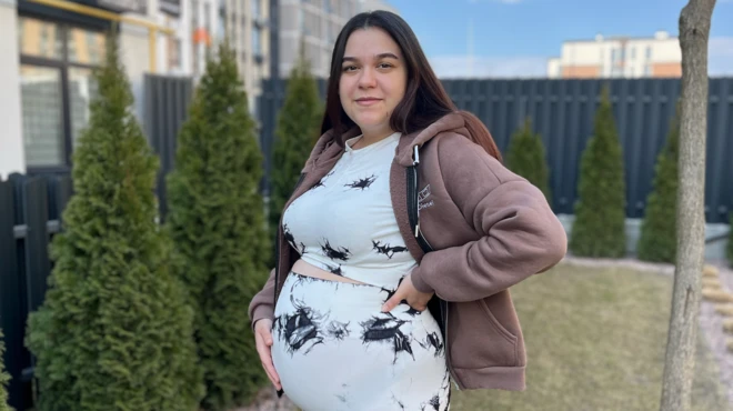 Una mujer embarazada está de pie en un jardín con la mano sobre el vientre. Tiene el pelo largo, liso y oscuro, y lleva un conjunto de blusa y pantalón blancos con detalles negros. También lleva una sudadera con capucha de color marrón claro.