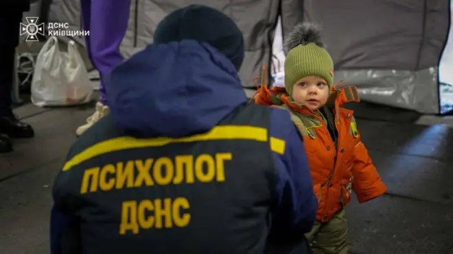 Человек в куртке с надписью "Психолог ДСНС" на спине присел на корточки перед ребенком в зимней одежде
