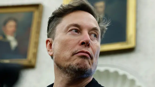 Elon Musk is shown in the Oval Office, looking away from the camera to the right against a blurred backdrop showing paintings of former US presidents on the wall behind him.