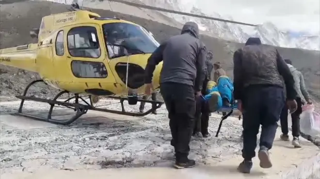 高山で、青い防寒着を着た人物が担架に乗せられ、黄色いヘリコプターに運ばれている