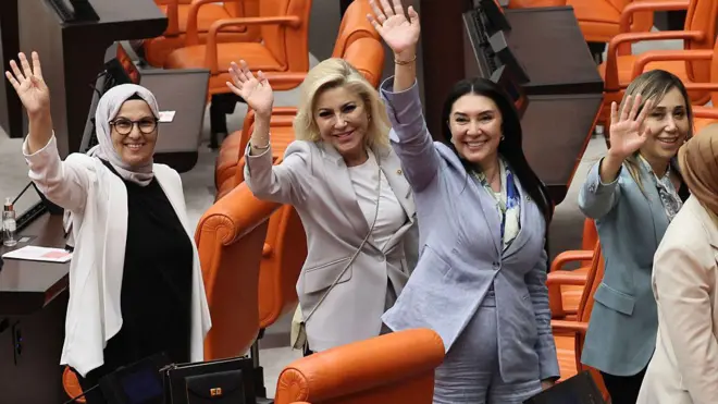 Biri başörtülü dört kadın milletvekili bir yere bakarak sağ ellerini selam verir gibi kaldırmış durumda. Meclis'teki turuncu renkli koltukların arasında ilerliyorlar