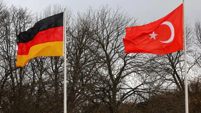 Türkiye ve Almanya bayrakları yanyana gönderde arka planda yapraksız bir ağaç var. 