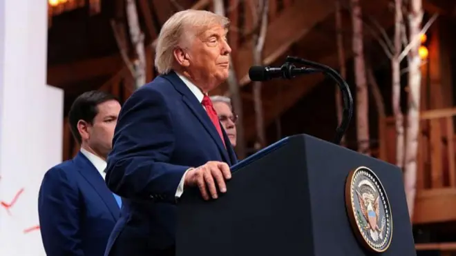Trump yandan görünüyor, kürsüde konuşuyor, gerisinde Marco Rubio'nun yarısı görünüyor.