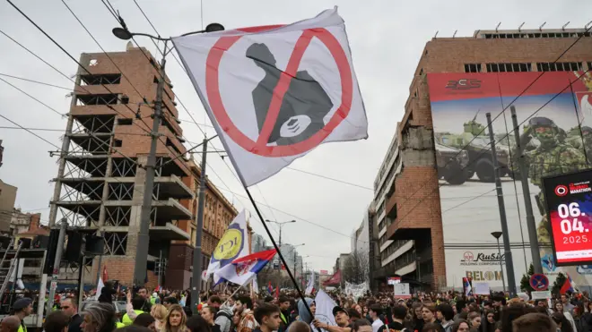 Группа молодых людей протестует с плакатами (в центре плакат с перечеркнутой фигурой) рядом со зданиями, поврежденными бомбардировками.