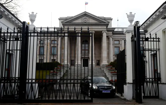Ambasada Rosji w Warszawie. Duży neoklasyczny budynek, do którego prowadzi ciężka żelazna brama. Na zdjęciu z otwartej bramy wyjeżdża czarny samochód z niebieską tablicą rejestracyjną.