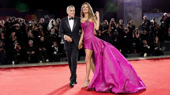 Smokin giymiş George Clooney ve pembe elbiseli Amal Clooney kırmızı halıda, arkada fotoğrafçılar