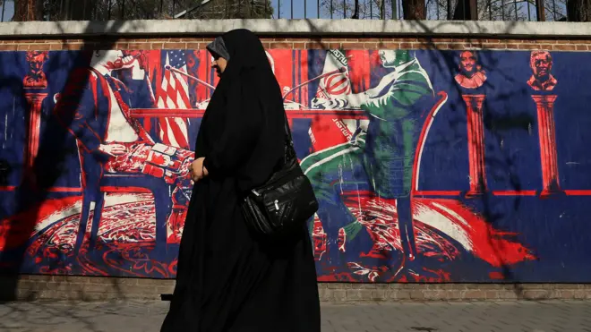A woman wearing a dark hijab and black dress walks past a mural on a wall depicting politicians from the US and Iran sitting opposite each other, taken in Tehran on 28 January.