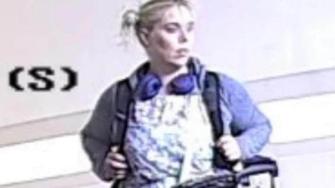 Footage captures Betro possibly in a shop with baggage. She has headphones on over a blue jacket and patterned dress.