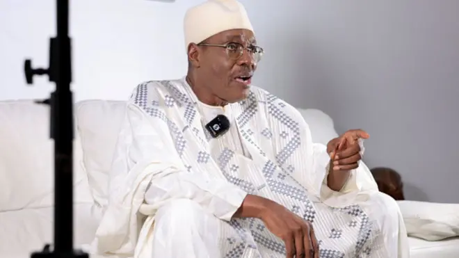 Tchiroma Bakary wear white traditional attire, siddon for chair dey tok to tori pipo