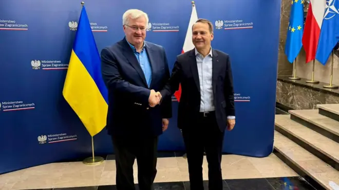 Сибіга та Сікорський тиснуть один одному руки під час офіційної зустрічі. На задньому плані видно прапори України, Польщі, Європейського Союзу та НАТО, а також стенди з логотипом польського міністерства закордонних справ.