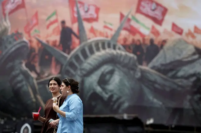 فتاتان تمران أمام لوحة إعلانية مناهضة للولايات المتحدة، تجسد تمثال الحرية ساقطاً وخلفه أعلام إيرانية ورايات كُتب عليها "يا لثارات الحسين"، في أحد شوارع طهران، إيران، في 3 أغسطس/آب 2026