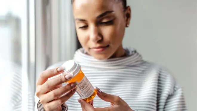 A young women with a stripey top holds a bottle of pills and reads the label.