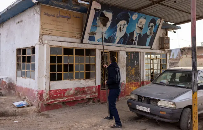 کارگری پوستر حسن نصرالله، حافظ اسد و علی خامنهای را در روز ۱۱ دسامبر در نزدیکی حلب پایین می کشد