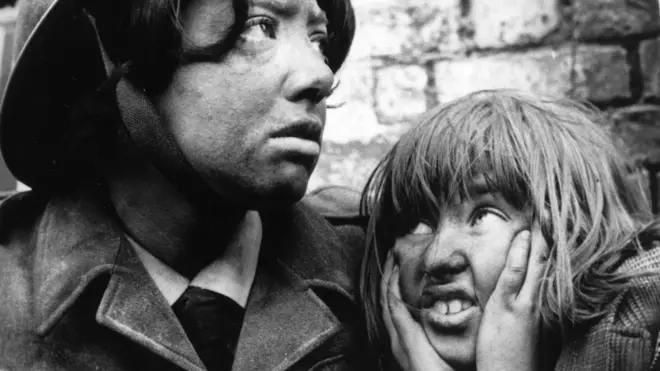 Црно бела фотографија на којој су млада жена у униформи и девојчица која рукама држи уши затворене. Лица обе делују прљаво. 