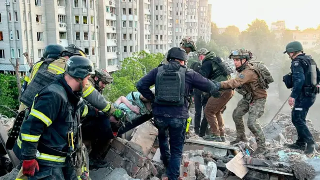 Група рятувальників і військових працює серед завалів зруйнованого будинку: вони спільно виносять людину на ношах із-під уламків. Довкола — купи бетону, цегли й металу, на тлі видно багатоповерхівки.