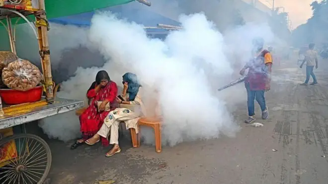 মশা নিয়ন্ত্রণে ফগার মেশিন দিয়ে ধোঁয়া ছাড়ছেন একজন কর্মী, সামনে একটি ভাসমান খাবারের দোকানের পাশে একটি বেঞ্চে বসে আছেন দুইজন নারী