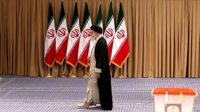 আয়াতুল্লাহ আলী খামেনি, পেছনে ইরানের সাতটি পতাকা। সামনে একটি ভোটের বাক্স।