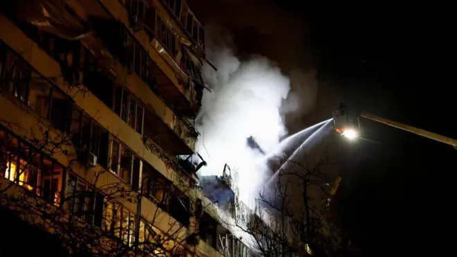 夜中に複数階の集合住宅へはしご車で消防士が消火剤をかけ、白煙が夜空に上がっている。破壊された建物の奥はオレンジ色に光っている