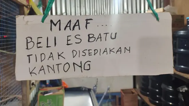 Sebuah poster tulisan manual di sebuah warung menunjukkan kepada pembeli bahwa pedagang tidak menyediakan kantong plastik untuk pembelian es batu. Poster ini bergantung dengan latar belakang plastik galon air mineral.