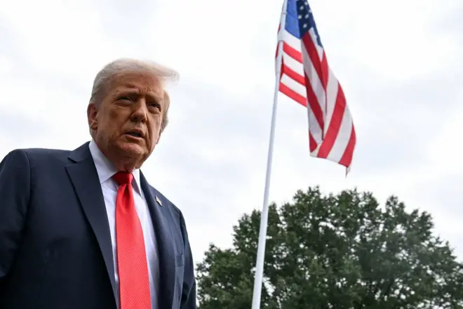 Trump em pé, com olhar sério, ao lado de bandeira dos EUA em área externa